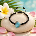 Pulsera de macramé con corazón de larimar sobre piedra clara con flor de frangipani, hoja de palma y pétalos