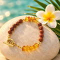 Pulsera de arena dorada y ámbar con baño de oro sobre piedra clara con flor de frangipani y hoja de palma