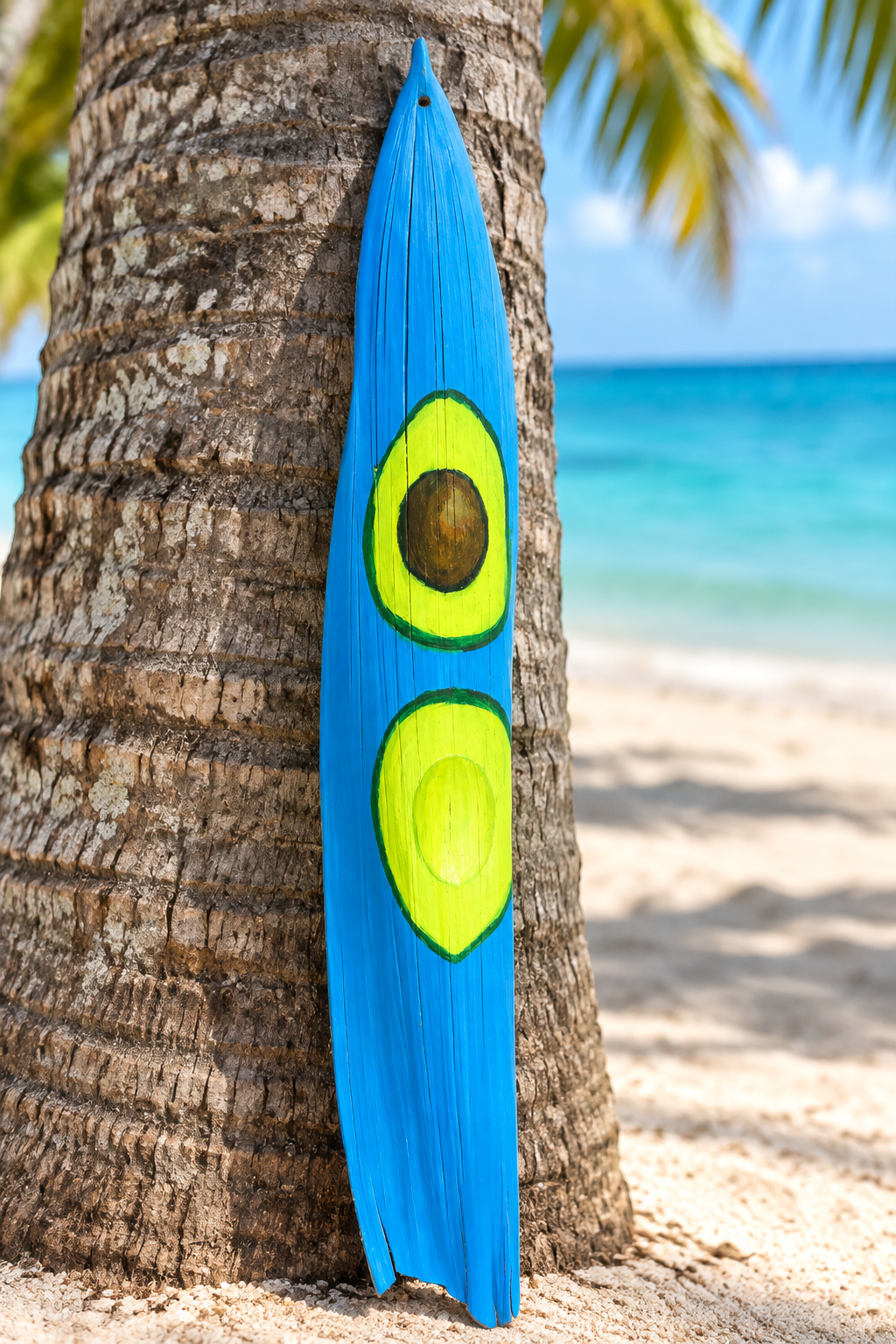 Penca de coco reciclada pintada a mano con diseño de aguacate, apoyada en el tronco de una palma frente al mar turquesa