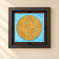 Cuadro decorativo de metal con motivo circular dorado sobre fondo azul, enmarcado en un ambiente luminoso de pared crema.