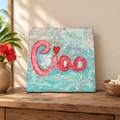 Cuadro de madera pintado con acrílicos con la palabra Ciao, apoyado sobre un mueble de madera