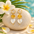 Aretes de ámbar con corazón de madreperla sobre piedra clara con flor de frangipani, hoja de palma y pétalos