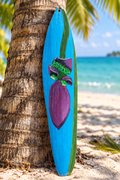 Penca de coco reciclada pintada a mano con diseño de flor de plátano, apoyada en el tronco de una palma frente al mar turquesa