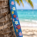 Penca de coco reciclada pintada a mano con diseño de flores tropicales, apoyada en una palma de coco frente al mar turquesa
